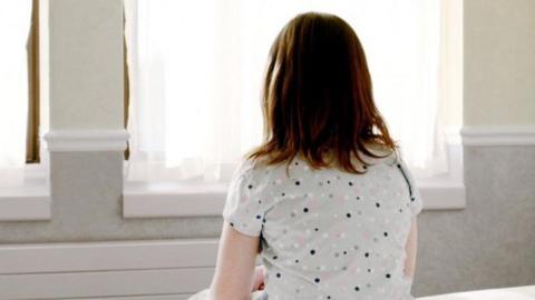 A girl with mid length brown hair is sat on a bed facing a window. She is wearing a grey top with black, pink and white dots on it