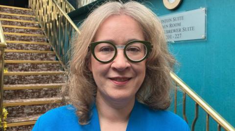 Selina Horshi has shoulder-length blonde hair and round glasses and is standing in front of a staircase which has a brown coloured carpet and brass bannisters. She is wearing a dark blue top. Over her left shoulder a teal coloured wall can be seen.