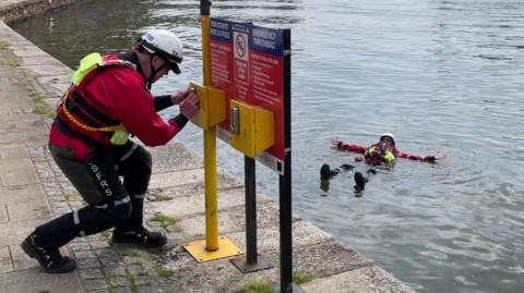 A person in red and black outfit and white hat handles a yellow box on a sign while another person floats in a river.