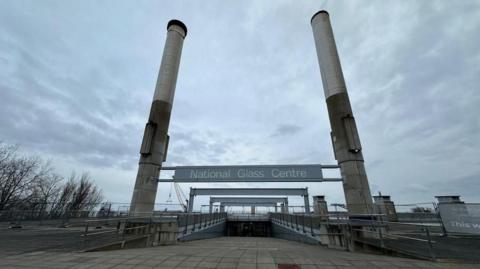The entrance to the National Glass Centre. The concrete path leads down a ramp. Meanwhile, two concrete pillars border either side. A grey sign reads: "National Glass Centre".
