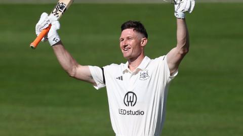 Cameron Bancroft holding a bat in the air in one hand and his helmet in the other and smiling while standing on the pitch