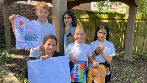 Image shows five children aged 11 wearing white polo shirts and holding things. Two of them are holding pencils, two are holding posters and one girl is holding a FIFA World Cup 2026 sticker book as well as a paper mache lion head mask.