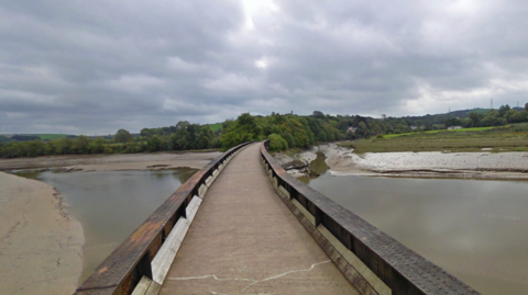 A narrow pedestrian bridge with wooden railings spans a muddy river or estuary. The sky is overcast with grey clouds, and trees and greenery line both sides of the water. No people are present in the scene.