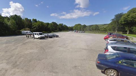 A large car park with loads of cars in it. Trees are around it.