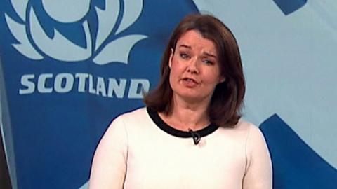 Woman in white top stands before large Scotland rugby flag during broadcast.