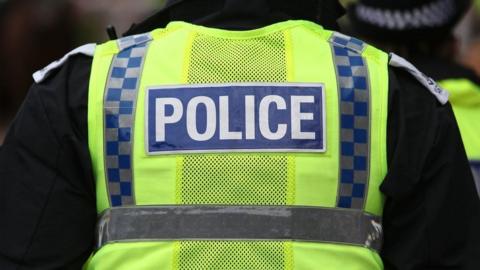 Police officer from behind wearing a yellow and blue high-visibility vest with the word "POLICE" across the back.