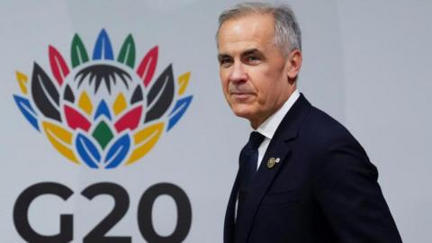 Prime Minister Mark Carney walks by the G20 logo against a white background. He has a slight smile on his face and wears a dark suit and tie. 