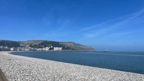 View of curved white pebble beach wrapped around sea, white buildings and hills in distance under a blue sky