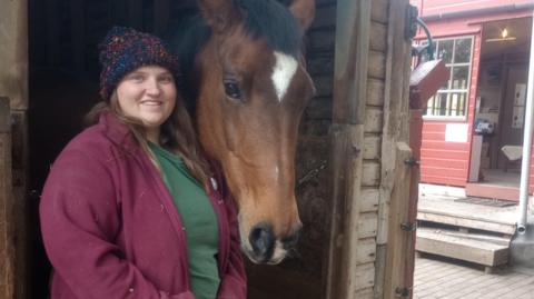 A young woman stands to the left of a horse, smiling at the camera. She wears a forest green t-shirt, open red zip fleece, and a big navy blue hat speckled with pink. The brown horse with a white forehead stripe and black mane holds its head close to the woman's chest, looking at the camera.
