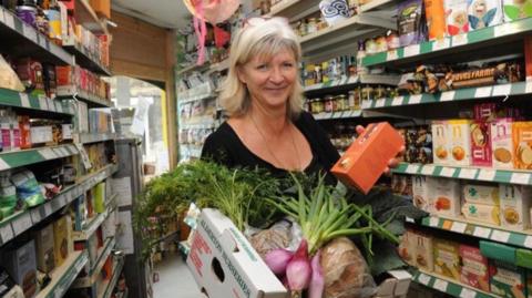 Chris Giles, a woman with a blonde flicked out bob, is holding an open cardboard box of food including large purple shallots, and what look like green leafy carrot tops, along with a loaf of bread. She is smiling at the camera warmly. Around her are shelves filled with produce including boxes of crackers and jars.