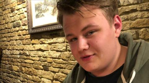 Harry Dunn wears a green hoody. He sits in front of a stone wall with a framed picture on it.