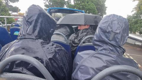People sat on the top of an open-top double decker bus in the rain. All are wearing waterproof coats mostly hidden by hoods. Two people also shelter under an umbrella pulled down over their heads and one man is looking back over his shoulder.