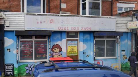 The outside of Daffodils Nursery in Smethwick. The walls are painted in a mural showing blue sky and flowers and a character standing by the door. There is a faded sign above the entrance in Waterloo Road.