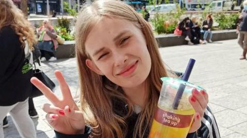 Image of Rhianan. She is holding up a bubble tea and is smiling with a peace sign for the camera. 