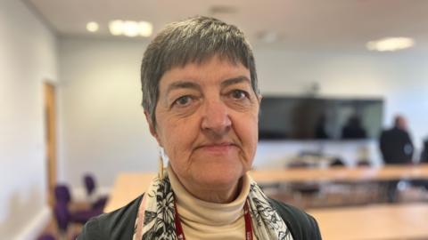 A woman with short, grey hair is pictured wearing a cream jumper with a white and grey scarf and a grey jacket. She is stood in an office environment but the background has been blurred. 