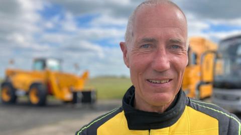 A man looks towards the camera wearing a yellow and black driving suit with the "JCB" logo on the front.