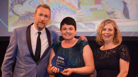 Man in suit and two women in sparkly dresses, one holding a tes award on stage 