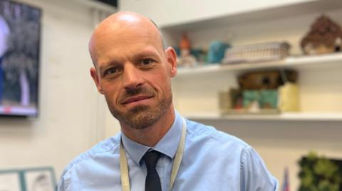 A man in his 30's with a bald head and slight facial hair wearing a blue shirt with a navy blue tie