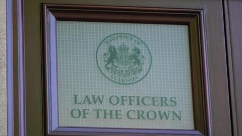 Photo of a brown door, with a white covered window. The text on the window says "law officers of the crown" and displays the Bailiwick of Guernsey crest.