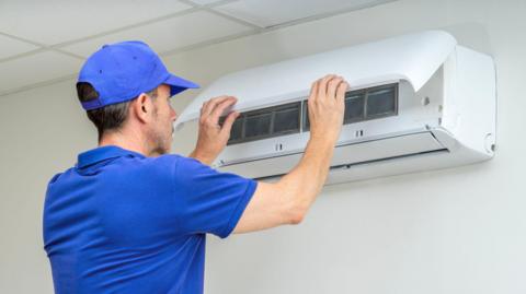 A man in blue t-shirt and a cap installing an air conditioning unit. They are a holding a panel and working on it whilst nailing it to the wall.