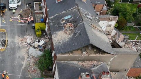 An aerial photo of a destroyed house following an explosion. Workers wearing hi vis are visible as well as a white van.