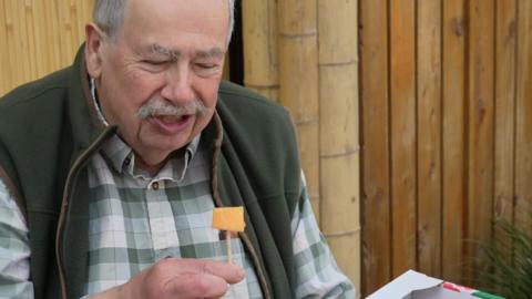 A man dressed in a shirt and overcoat holds a melon on a stick to taste test it