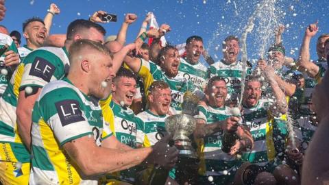 About 20 rugby players in green and white hooped tops hold a cup and pop champagne as they celebrate a win. Their mouths are open as if they are cheering and some of their arms are in the air.