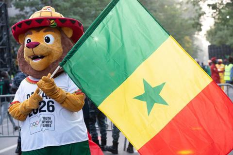 This image shows a friendly-looking lion mascot with a soft, golden-brown face, large expressive eyes, and a wide, cheerful smile. It wears a distinctive, oversized conical hat (a traditional Saloum hat) featuring yellow and orange patterns with a red brim. The mascot is dressed in a white t-shirt that prominently displays the text "DAKAR 2026" above the official Youth Olympic Games logo and holds the national flag of Senegal (red, yellow, and green) in a celebration parade.