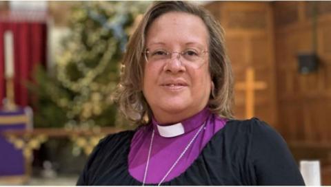 Closeup of Rt Rev Tricia Hillas wearing a dog collar with a purple shirt under a black dress. There is a large Christmas tree in the background.