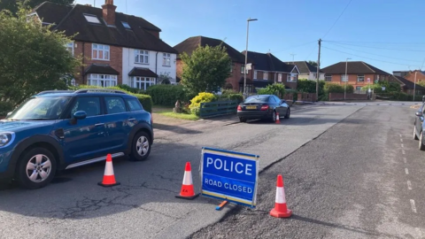 A police cordon in a residential street with two cars parked on the left hand side and one on the right hand side 