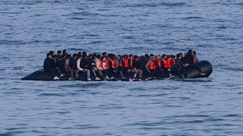 A black inflatable boat full of people - some wearing red life jackets - crossing the English Channel in June.