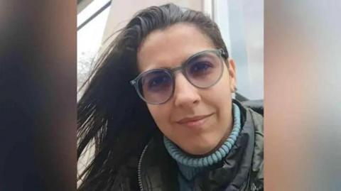 A picture shows the face of Barreto. She is wearing a blue turtleneck under a coat and glasses.