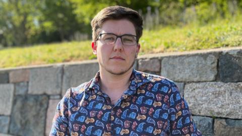 Sam is wearing glasses and a patterned button-up shirt sits on a wooden bench beside a stone wall in a sunny park, with green grass and trees in the background.