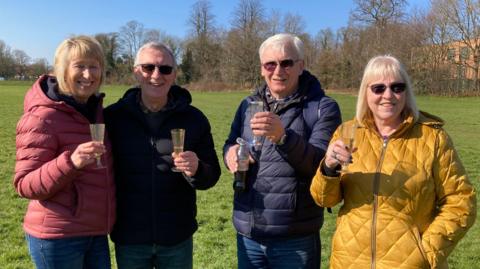 Two men and two women standing in a green field lined by trees. The men are wearing dark anoraks, while one woman has a red anorak and the other is wearing yellow.  They are holding plastic champagne glasses.