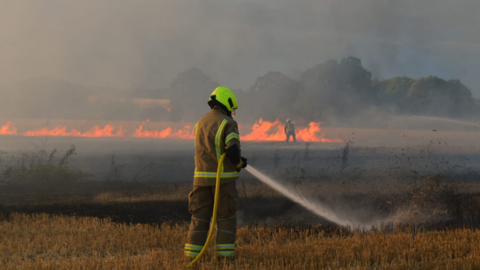 A firefighter with his back to the camera trains a hose on a smouldering field. In the distance, beyond him, another firefighter is tackling a line of flames. The air is thick with smoke.