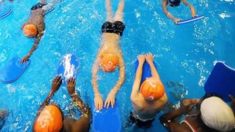 Children in a pool taking part in a swimming lesson. They are swimming on their fronts, holding out blue floats, while adults stand in the water watching.