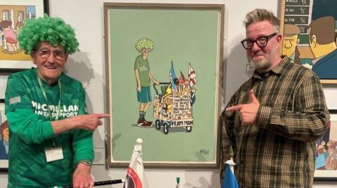 Fundraiser John Burkhill is standing on the left of the picture with his pram in front of him pointing to a painting of him which is being raffled off. On the right is the Sheffield artist Pete McKee, he is wearing brown trousers, and a dark-green checked shirt. He has short light-brown hair and is wearing dark-rimmed glasses.