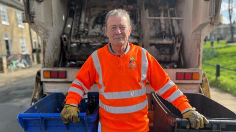 A refuse collector dressed in orange with his hands on two wheelie bins either side of him, one blue, one black. Behind him is a bin lorry