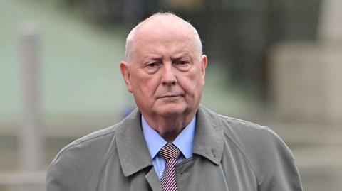 A man with white balding hair is pictured walking towards the camera. He is wearing a grey coloured trench coat, a blue shirt and a striped tie. The background is blurred. 