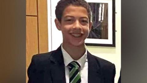 Noah Donohoe, a boy with dark hair, wearing a black blazer, white shirt and green, black and white tie.