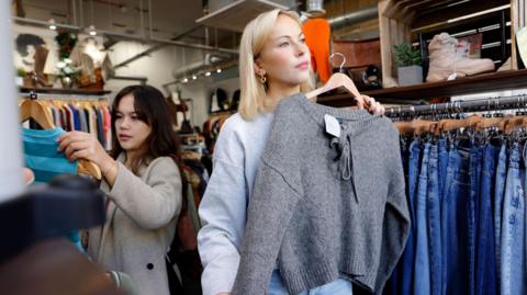 Blonde woman holds up grey cardigan in clothes shop whilst another brunette woman looks at a dress behind her.