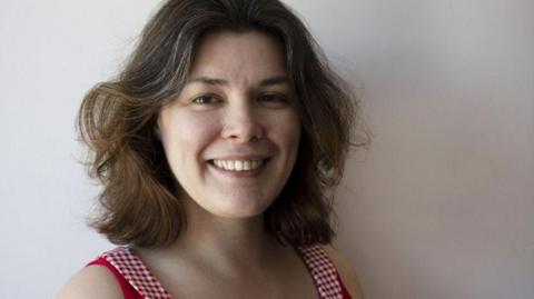 A woman with dark brown hair smiles broadly as she looks into the camera. She is wearing a red and white checked dress.