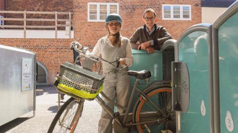 Councillor Hannah Cooper wearing a blue helmet holds a bike with a basket on the front next to three green and grey bike lockers. Chris Jardine, co-founder of Bikedok, is stood behind one of the lockers wearing a brown fleece and round glasses.