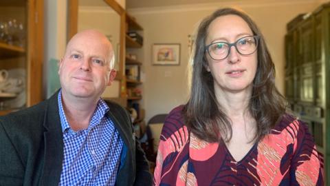 Darren and Jennifer looking at the camera. Darren is wearing a blue and white checked shirt under a dark grey jacket. Jennifer is wearing a red dress with orange, pink and navy patterns.