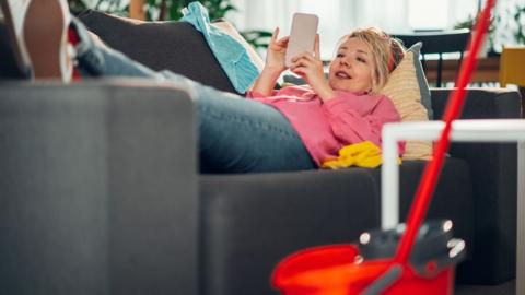 Woman lounging on a couch, engrossed in her smartphone, while a mop and bucket sit nearby, suggesting procrastination from cleaning tasks in a cozy home setting