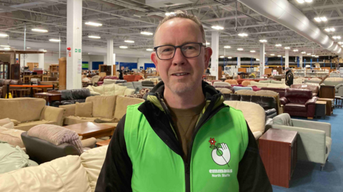 A man in glasses, wearing a green top with Emmaus North Staffs on the front stands in a very large warehouse type building. Behind him furniture stretches into the distance