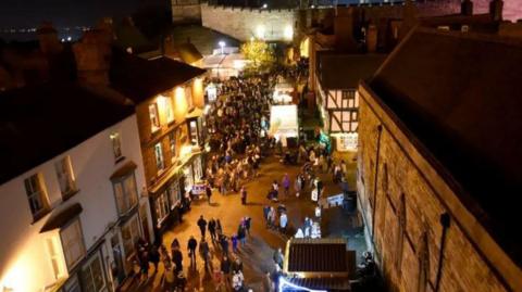 A bird's-eye view of the streets of Lincoln in the evening. There are crowds of people gathering around market stalls. The walls of Lincoln Castle are in the background