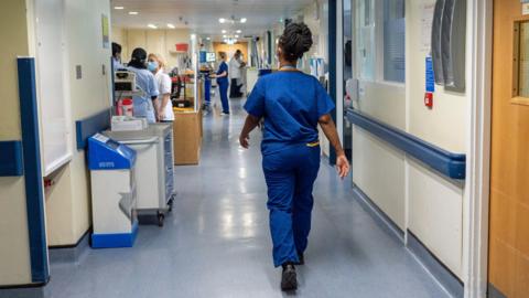 A nurse in blue overalls walks down a busy hospital coridoor