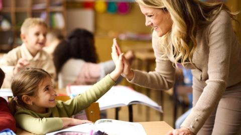 A young teacher is leaning over the desk of two under-10 pupils and is high-fiving the nearest child. Both are smiling.