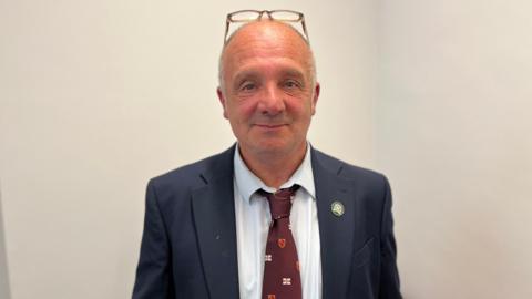 A bald man, with a pair of glasses on his head, looking at the camera with a slight smile. He is wearing a white shirt, a dark blue blazer and a burgundy tie with small Guernsey flags on it.